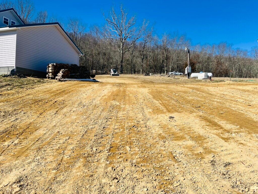 A rural dirt road with a house situated in the background, set against a backdrop of fields and trees.